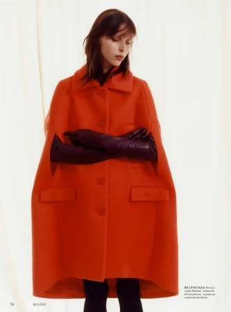 Model wearing a vibrant red coat and long dark gloves against a pale draped curtain background.