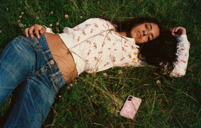 Young woman with long dark hair lying in tall green grass wearing denim jeans and a patterned zip hoodie.