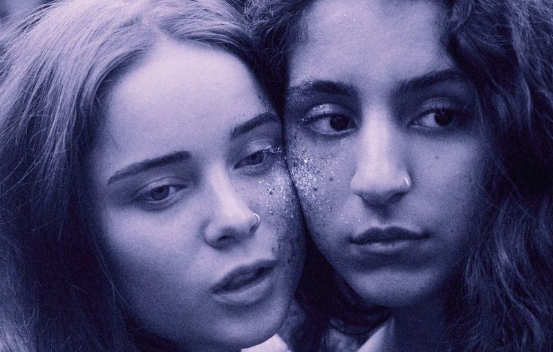 Close-up portrait of two young women with glittering makeup pressing cheeks together under purple monochrome lighting.