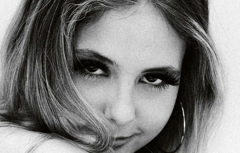 Close-up black and white portrait of a young woman with dark eye makeup and hoop earrings looking directly at the camera.