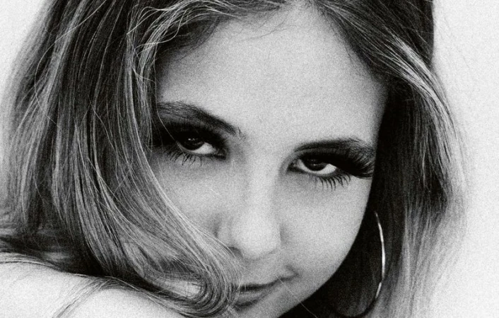 Close-up black and white portrait of a young woman with dark eye makeup and hoop earrings looking directly at the camera.