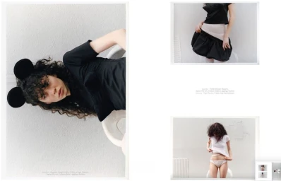 Model with dark curly hair wearing black mouse ear headband and black top against a pale grey textured wall background.