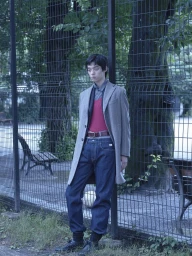 Young man leaning against a wire fence wearing a grey coat, red top, and dark denim jeans