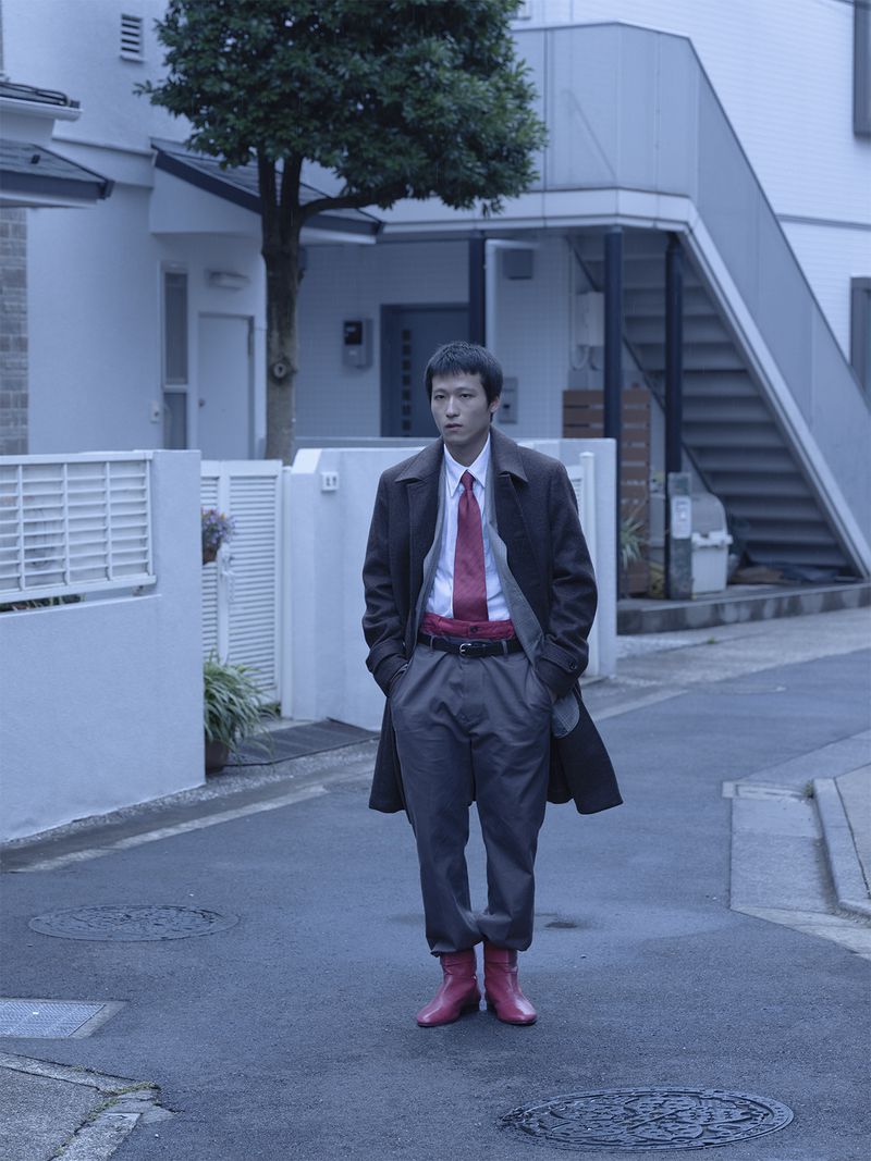 Man wearing a brown coat and red boots standing on a narrow street framed by residential buildings