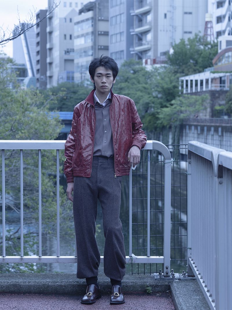 Young man leaning against a railing on an urban bridge wearing a red jacket and dark trousers