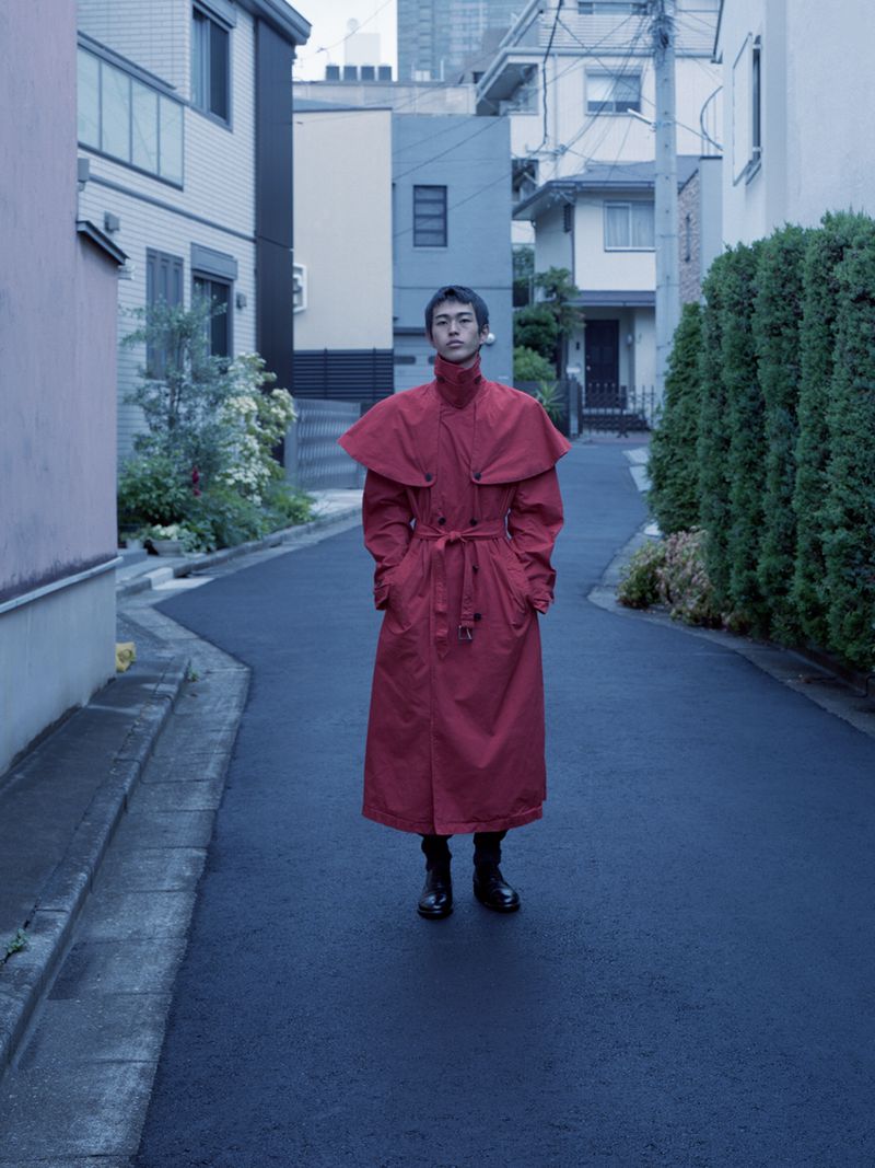 Man wearing a long red belted coat standing on a narrow residential street lined with houses and tall hedges