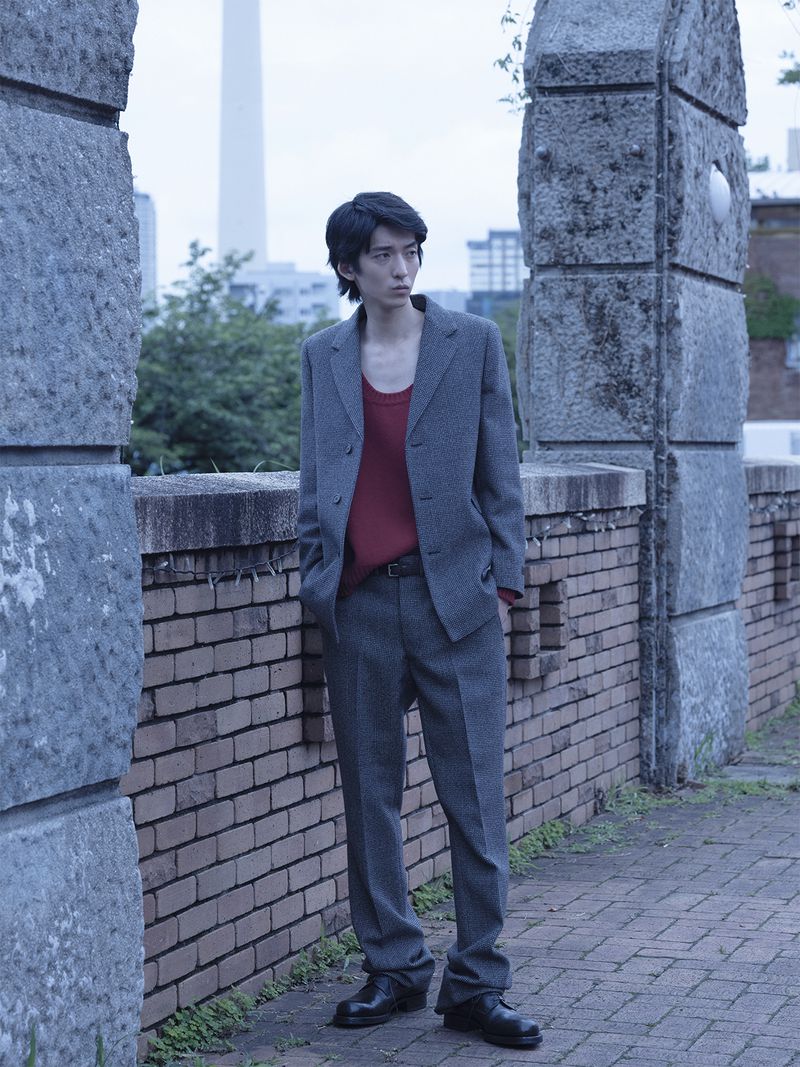 Young man in a grey suit jacket and trousers standing near a brick wall outdoors