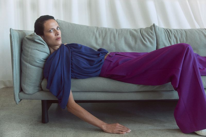 Aymeline Valade reclining on a grey sofa wearing a blue top and purple trousers for M Le magazine du Monde