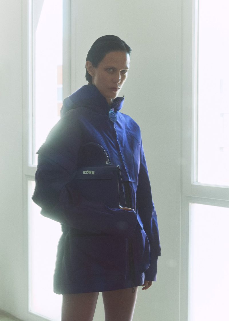 Model Aymeline Valade wearing a blue jacket and holding a handbag in front of bright windows for M Le magazine du Monde