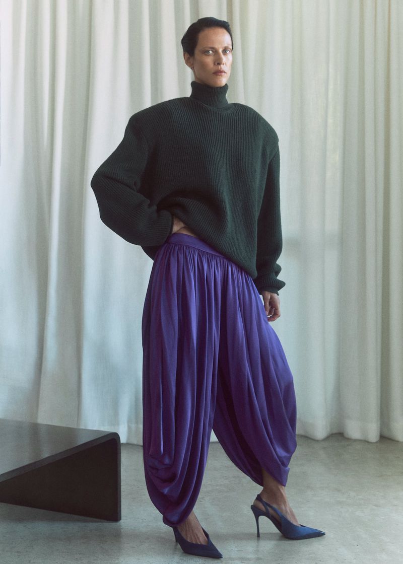 Model Aymeline Valade wearing a dark green turtleneck and purple trousers against white curtains for M Le magazine du Monde.