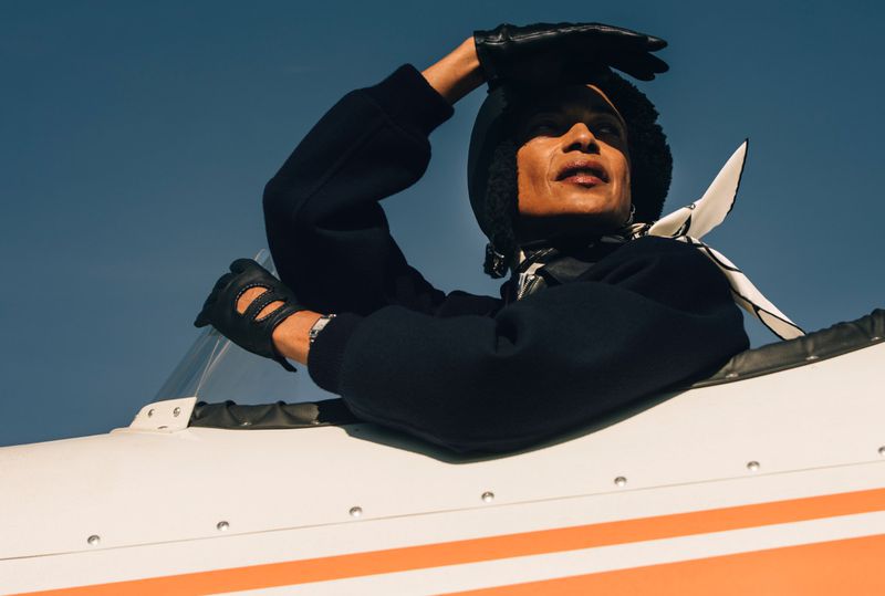 Model in aviator cap and leather gloves leaning out of an aircraft cockpit against a clear blue sky