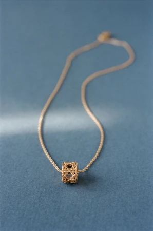Gold chain necklace with a textured cylindrical pendant resting on a textured blue background.