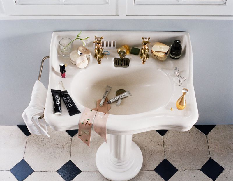 High-angle view of a vintage white porcelain pedestal sink cluttered with Dior cosmetic products, perfume bottles, and skincare items.