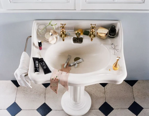 High-angle view of a vintage white porcelain pedestal sink cluttered with Dior cosmetic products, perfume bottles, and skincare items.