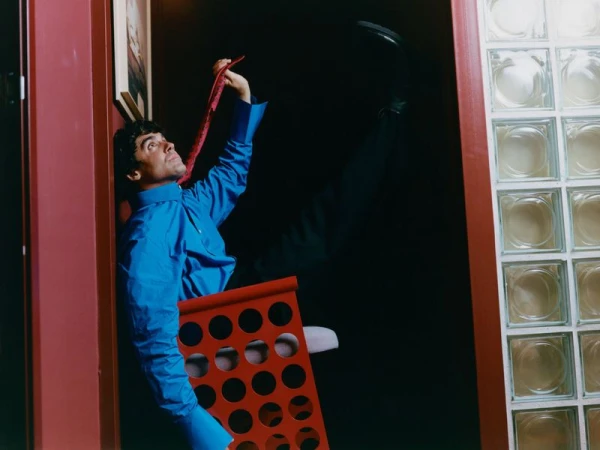 Man wearing a vibrant blue shirt holds a red tie while posing inside a narrow interior corridor next to a glass block wall.
