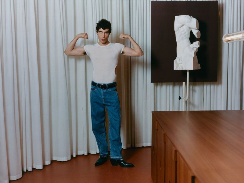 Man wearing a white t-shirt and blue jeans flexing his arms in an interior space with white curtains and a white sculptural bust