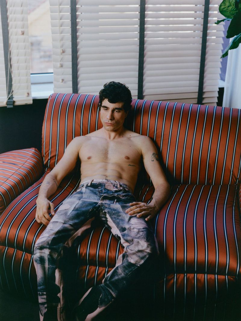 Shirtless man seated on a red and black striped sofa in front of a window with blinds