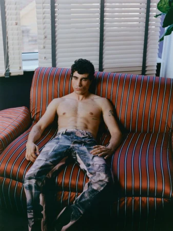 Shirtless man seated on a red and black striped sofa in front of a window with blinds