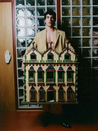 A man wearing an open tan blazer holding a detailed model house against a glass block wall.