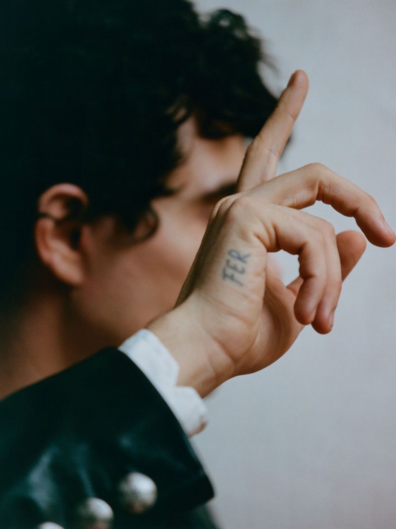Close-up portrait of a man holding his hand up with a small tattoo on his finger, wearing a dark jacket.