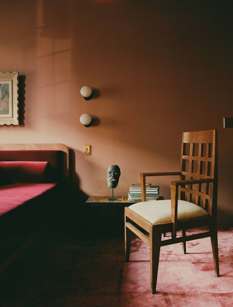 Interior view of a bedroom with terracotta walls, a wooden chair, red velvet bed, and a decorative sculpture on a small side table.