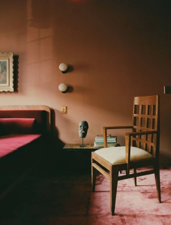 Interior view of a bedroom with terracotta walls, a wooden chair, red velvet bed, and a decorative sculpture on a small side table.