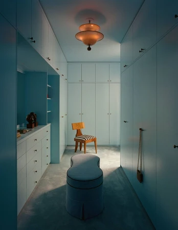 Interior view of a pale blue walk-in closet with built-in cabinetry, a central upholstered ottoman, and a wooden chair.