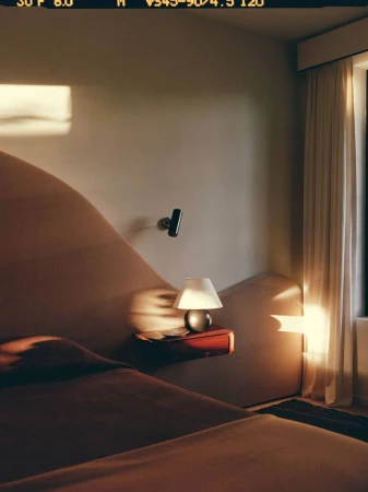 Interior bedroom view showing a low-profile bed with a sculpted curved headboard and warm evening sunlight falling across the wall.