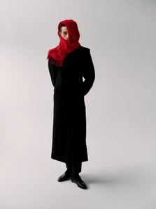 Model wearing a long black coat and a bright red scarf wrapped over the head against a light grey studio background.