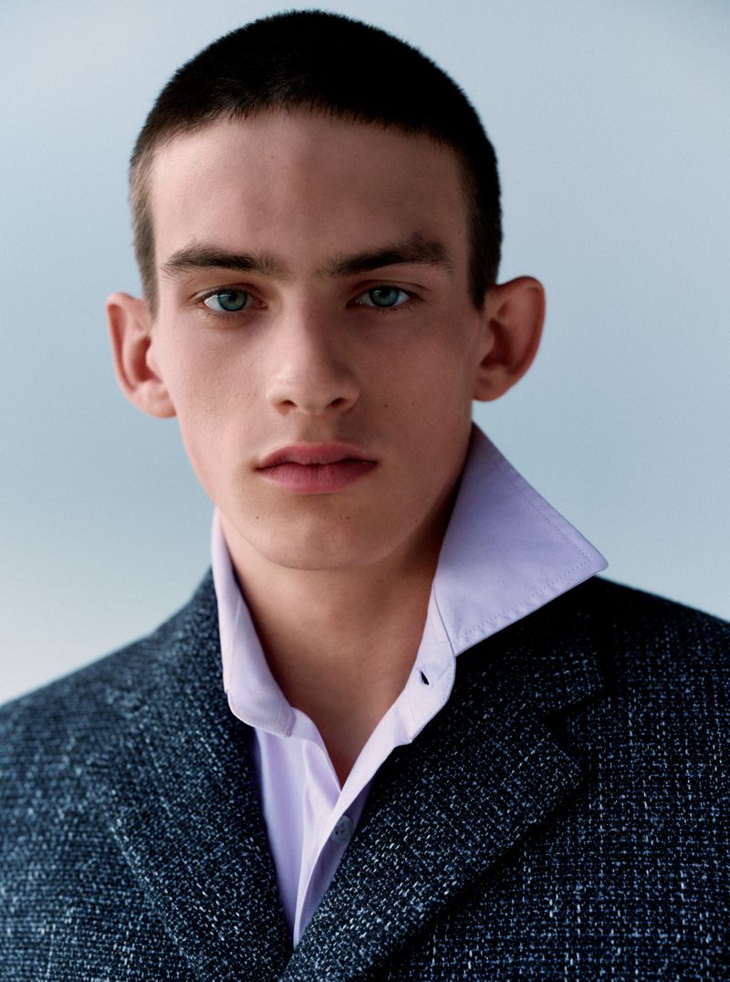 Close-up portrait of a young male model wearing a textured dark jacket and light purple shirt against a pale background