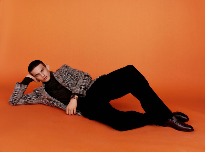 Model in plaid blazer and dark trousers reclining on an orange seamless studio background