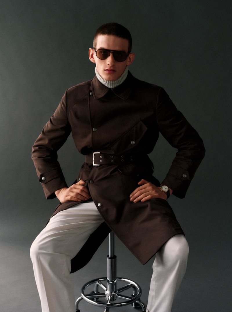 Model seated on a stool wearing a dark brown trench coat, white trousers, and sunglasses against a grey studio backdrop.
