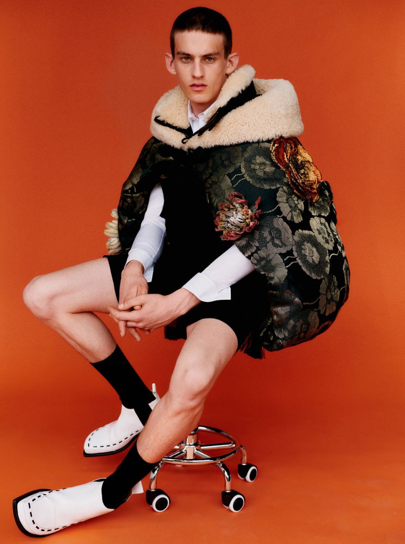 Model seated on a rolling stool wearing a patterned cape with shearling hood, shorts, and white boots against an orange background