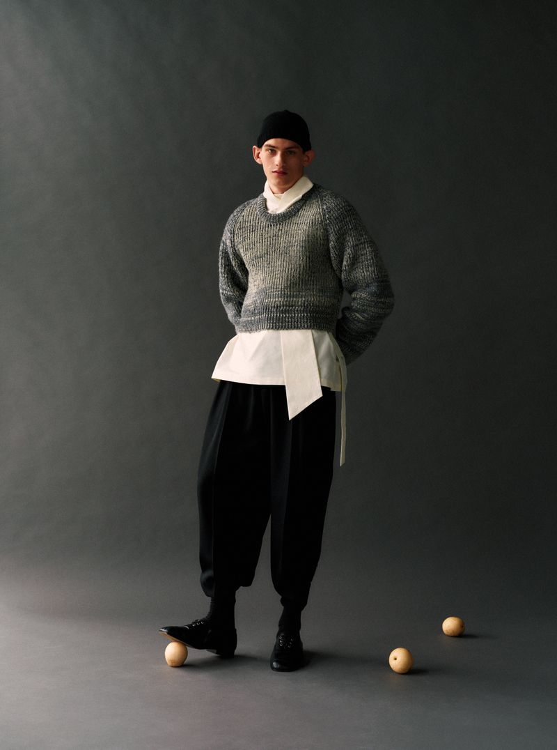 Model wearing a grey knit sweater and black trousers with a foot resting on a round fruit against a grey background.
