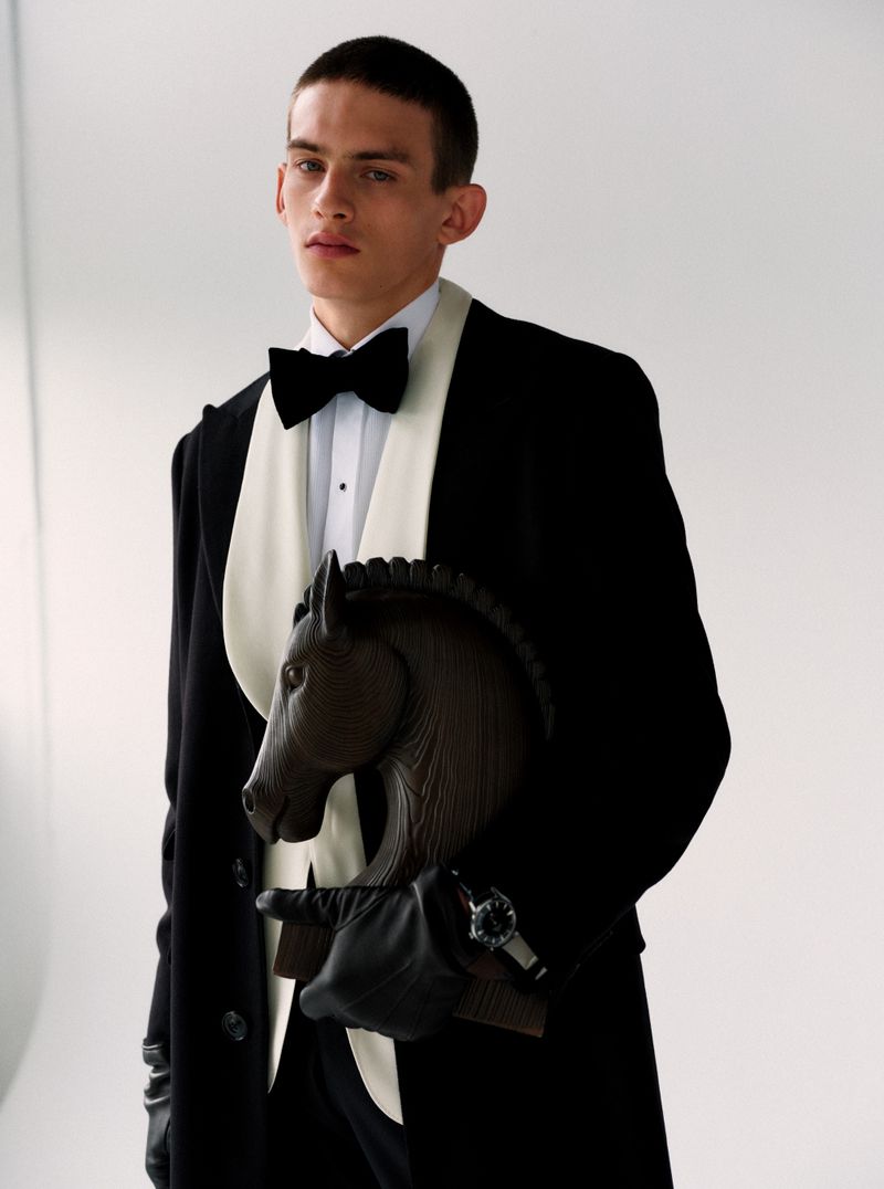 A male model wearing a tuxedo and black coat holding a dark horse figurine against a white seamless background.