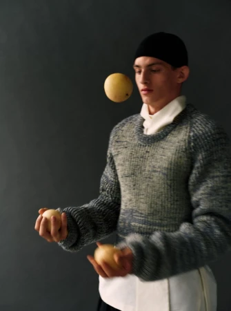 Young male model juggling yellow fruit while wearing a chunky knit sweater and black cap against dark studio background.