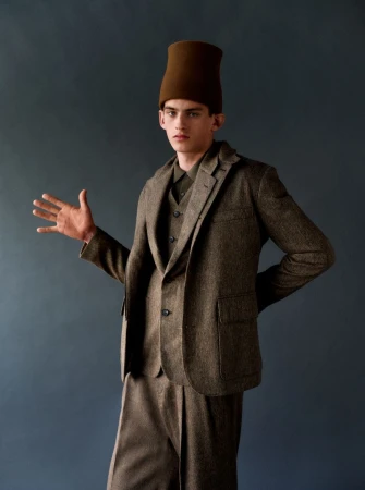 Model wearing a brown tweed suit and tall brown hat posing against a dark background