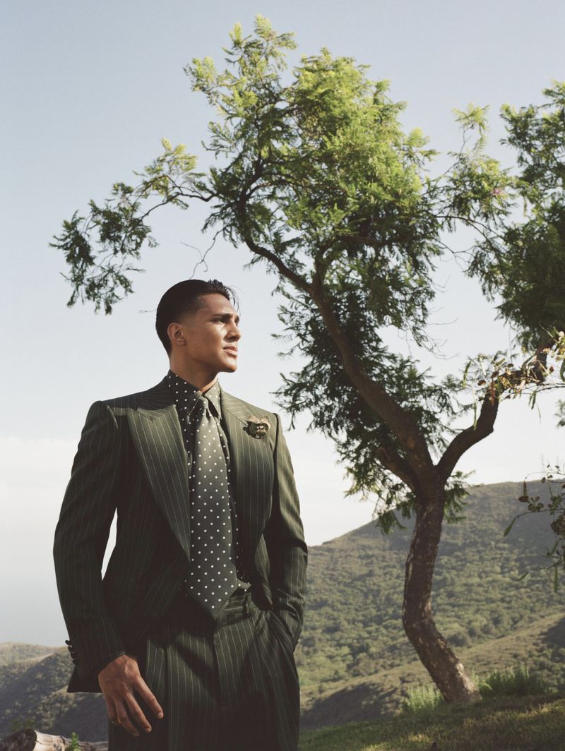 A man wearing an olive green pinstripe suit and polka dot tie stands outdoors near a tree with rolling hills in the background.