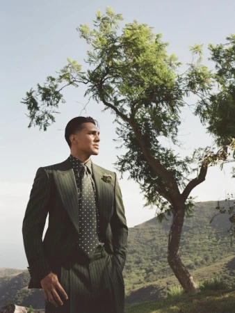 A man wearing an olive green pinstripe suit and polka dot tie stands outdoors near a tree with rolling hills in the background.