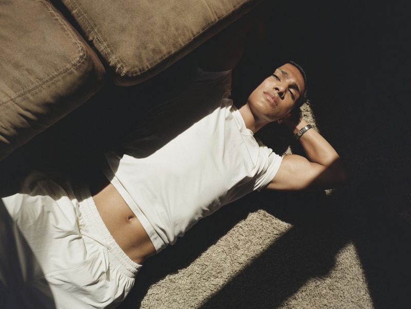 Man in white t-shirt lying on carpeted floor under bright sunlight next to a brown couch.