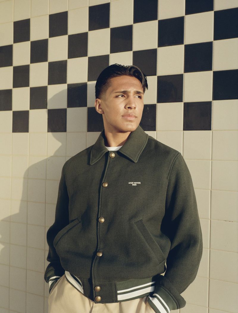 Young man wearing a dark green jacket and light trousers standing against a tiled wall with a checkerboard pattern.