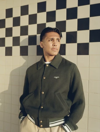 Young man wearing a dark green jacket and light trousers standing against a tiled wall with a checkerboard pattern.