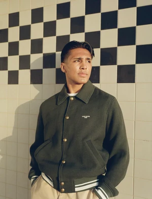 Young man wearing a dark green jacket and light trousers standing against a tiled wall with a checkerboard pattern.