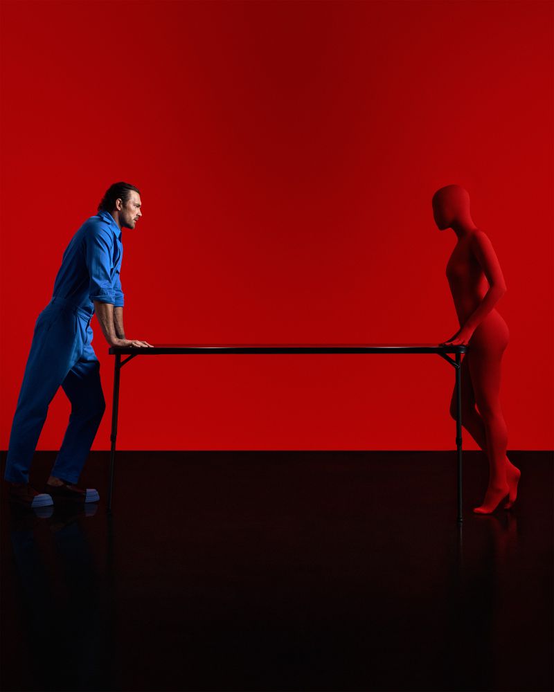 Man in blue jumpsuit standing opposite a figure in a red bodysuit across a black table against a vivid red backdrop.