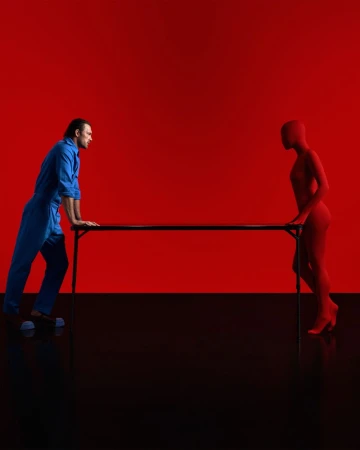 Man in blue jumpsuit standing opposite a figure in a red bodysuit across a black table against a vivid red backdrop.