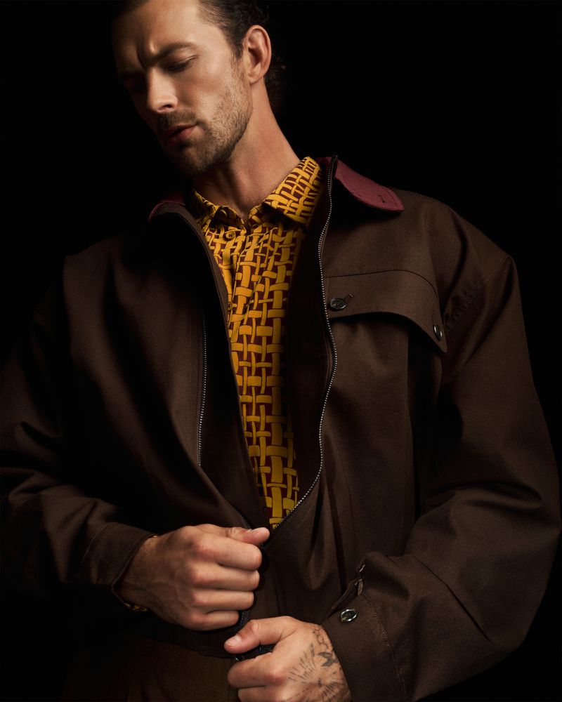 Christian Hogue in a brown jacket and patterned yellow shirt against a black background for TMRW Magazine editorial.