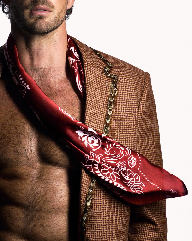 Close-up of male model Christian Hogue wearing a patterned red scarf and brown houndstooth jacket against white.