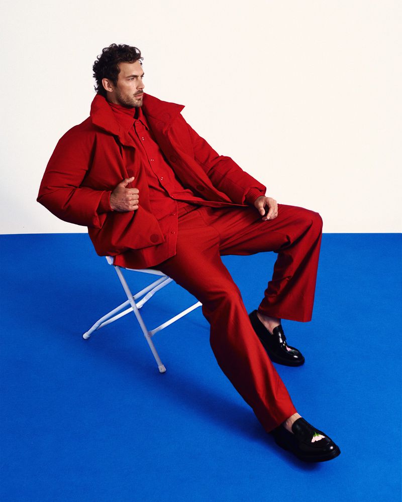 Christian Hogue wearing a vibrant red puffer jacket and matching trousers while seated on a white folding chair against a two-tone studio backdrop.