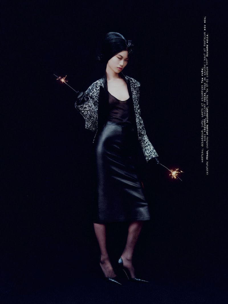 Model wearing a patterned jacket and black leather skirt holding sparklers against a black background.