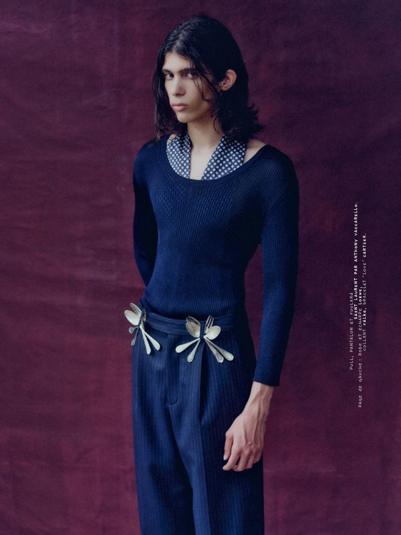 Model with long dark hair wearing a blue textured top and trousers with forks tucked into the waistband against a dark burgundy backdrop.